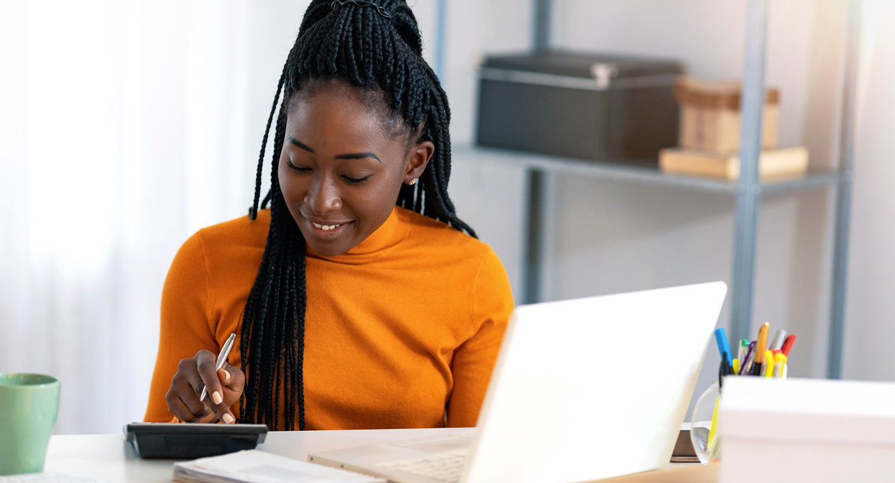 A Black woman with long hair in an office using a calculator and laptop, planning to pay either her credit card’s statement balance or current balance.