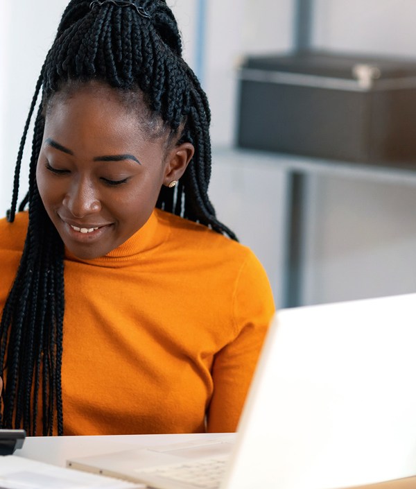 A Black woman with long hair in an office using a calculator and laptop, planning to pay either her credit card’s statement balance or current balance.