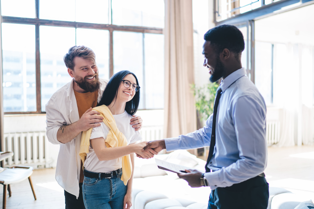 Image of black man holding clipboard and shaking hands with a white woman while her husband stands behind her.