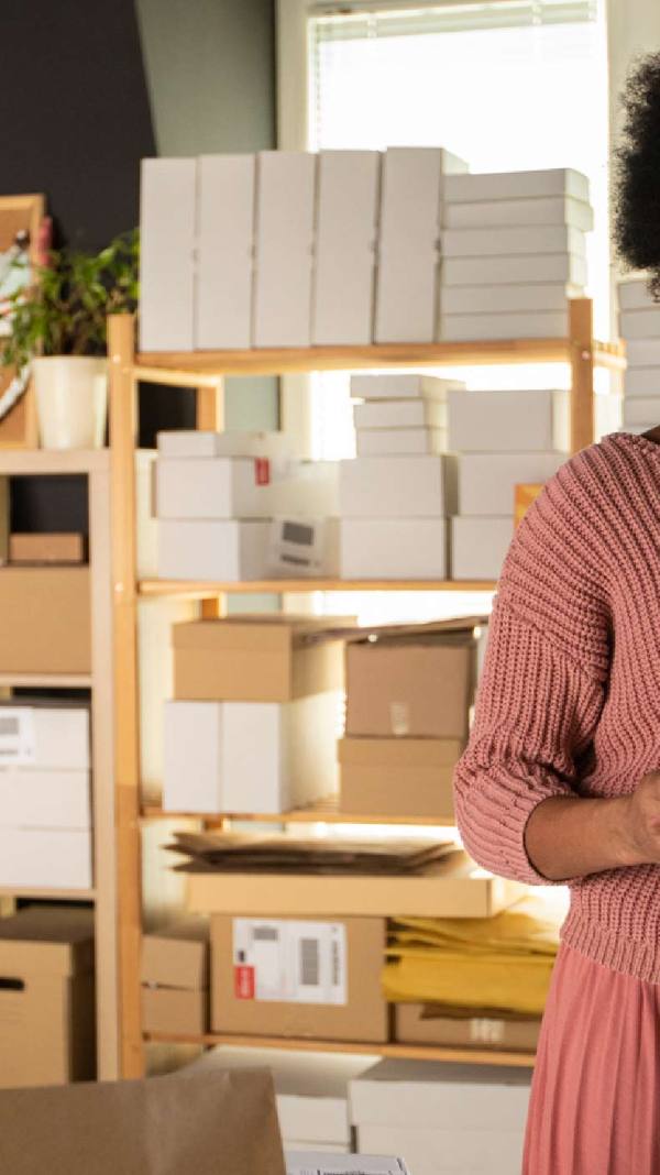 A Black woman and small business owner is in a shop surrounded by packages, looking at her tablet, indicating she might be using the cost of goods sold formula for her accounting.