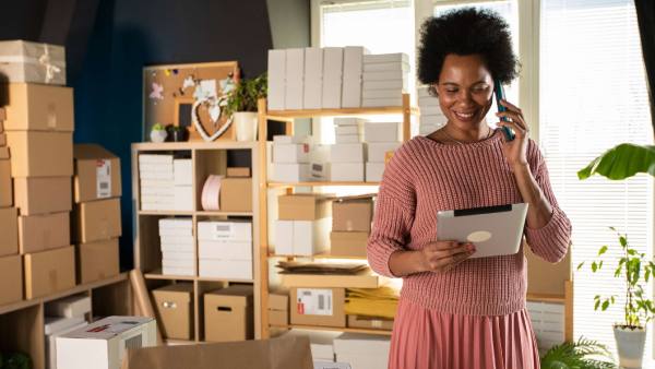 A Black woman and small business owner is in a shop surrounded by packages, looking at her tablet, indicating she might be using the cost of goods sold formula for her accounting.