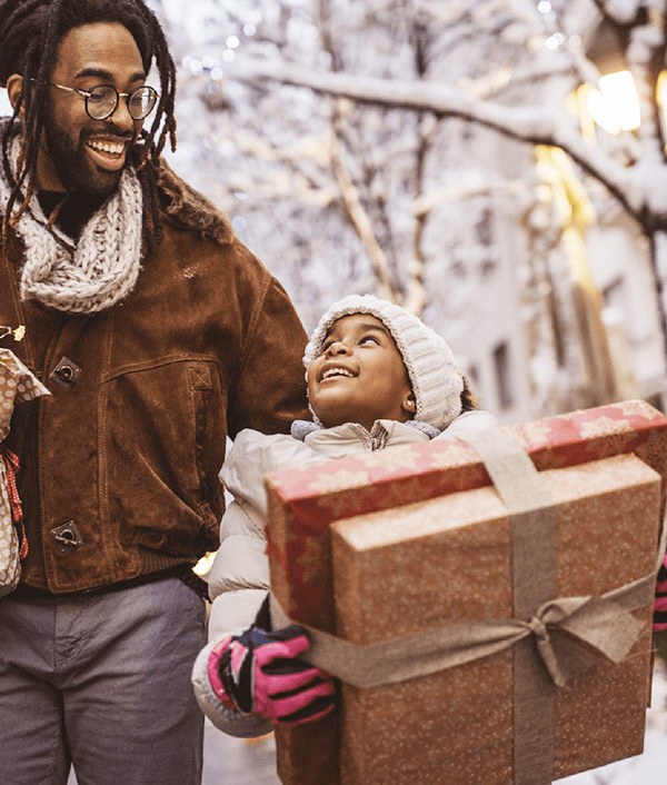 A Black man holds a small Christmas tree walking in a snowy city alongside a kid holding two gift boxes while smiling at each other, indicating they have their holiday budgeting in order.