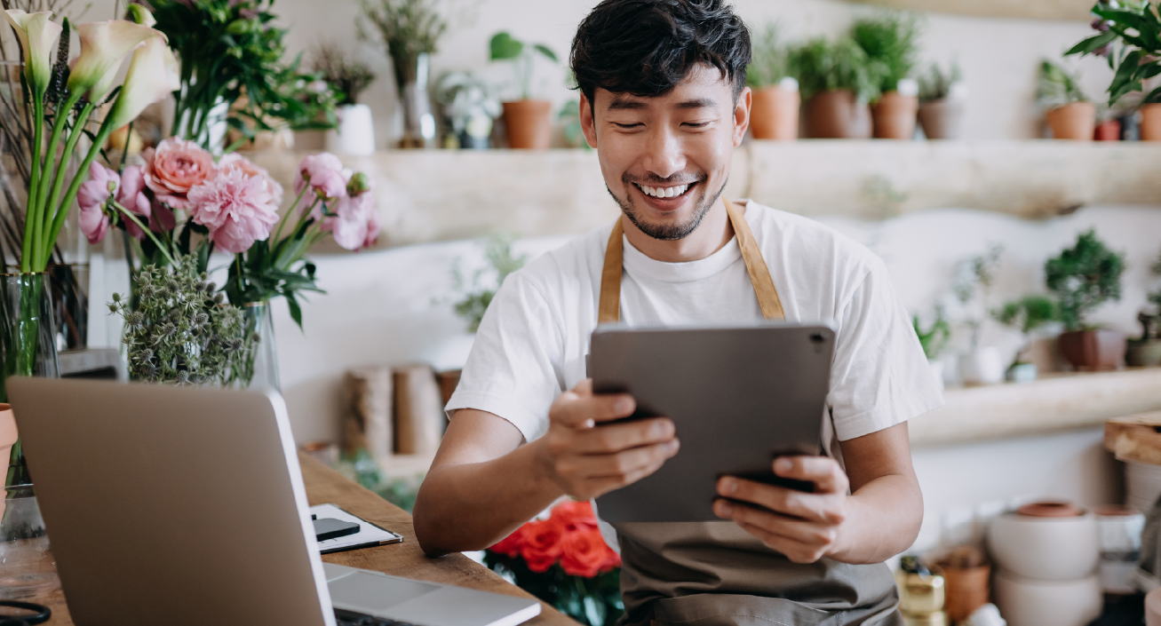 An Asian man and small business owner is in a flower shop, looking at his iPad, indicating he might be reviewing the entrepreneur salary he set for himself