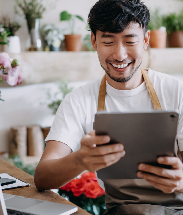 An Asian man and small business owner is in a flower shop, looking at his iPad, indicating he might be reviewing the entrepreneur salary he set for himself