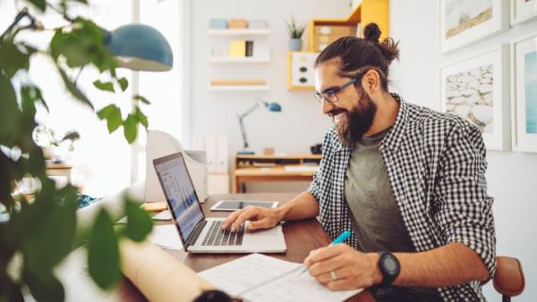 A man sits at a desk in front of his laptop, writes something down, indicating he is happy about the money he is saving from doing the 52-week money challenge.