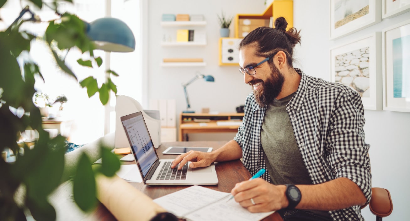 A man sits at a desk in front of his laptop, writes something down, indicating he is happy about the money he is saving from doing the 52-week money challenge.