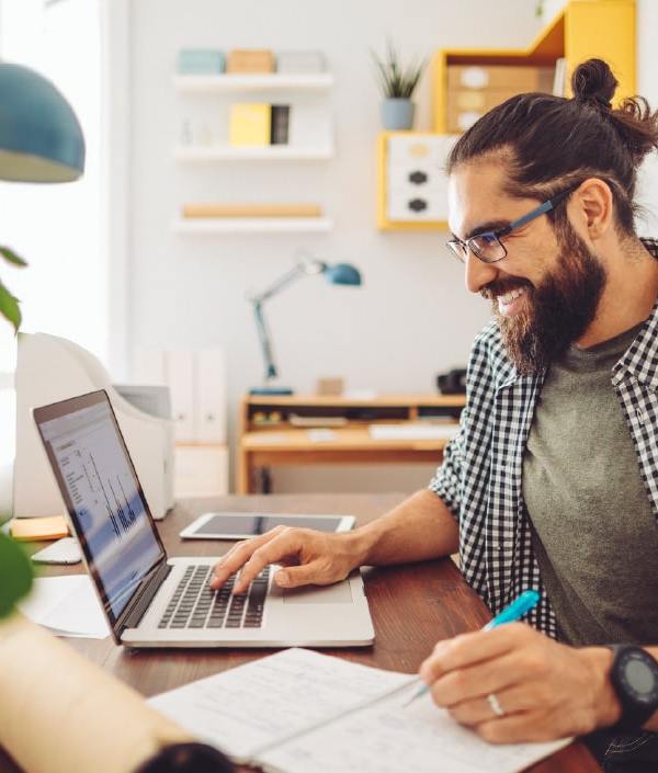 A man sits at a desk in front of his laptop, writes something down, indicating he is happy about the money he is saving from doing the 52-week money challenge.