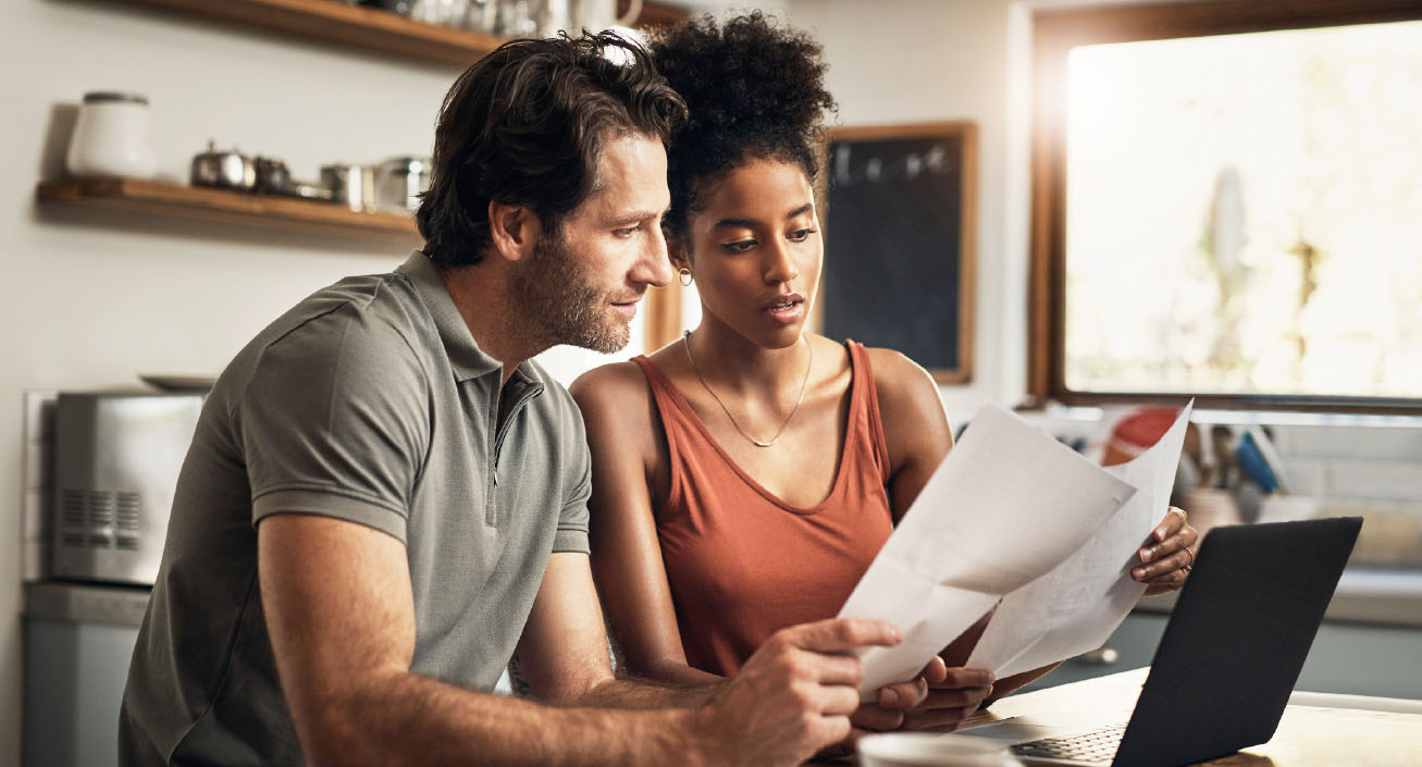 A couple sits at their kitchen table in front of their computer while looking at their bank statements, indicating they want to start having better money habits.