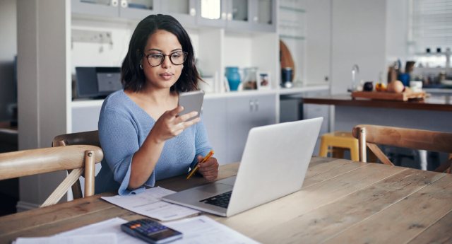 A woman sits at her kitchen table in front of her computer while staring at her phone, indicating she's using a budgeting app to develop better money habits.