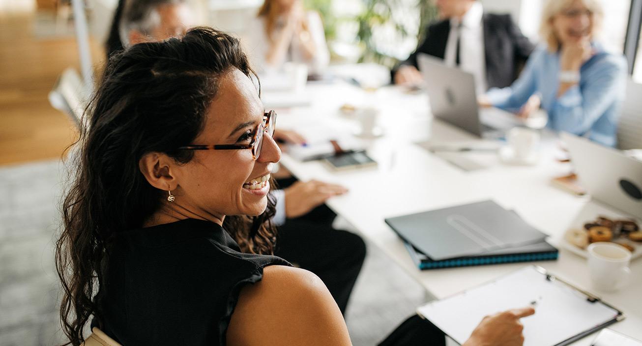 A woman sits at a work meeting and smiles at her cowokers, indicating she believes she has one of the most fulfilling jobs.