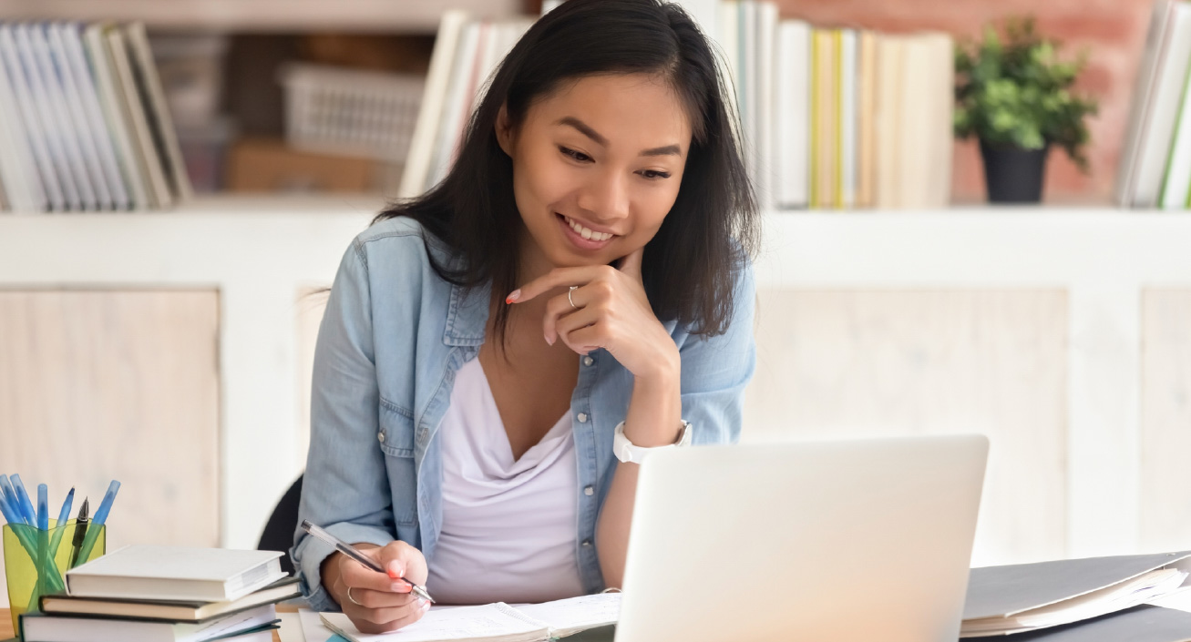 A woman sits in front of her laptop with a notebook open while taking notes, indicating she's learning how to write a first job resume.