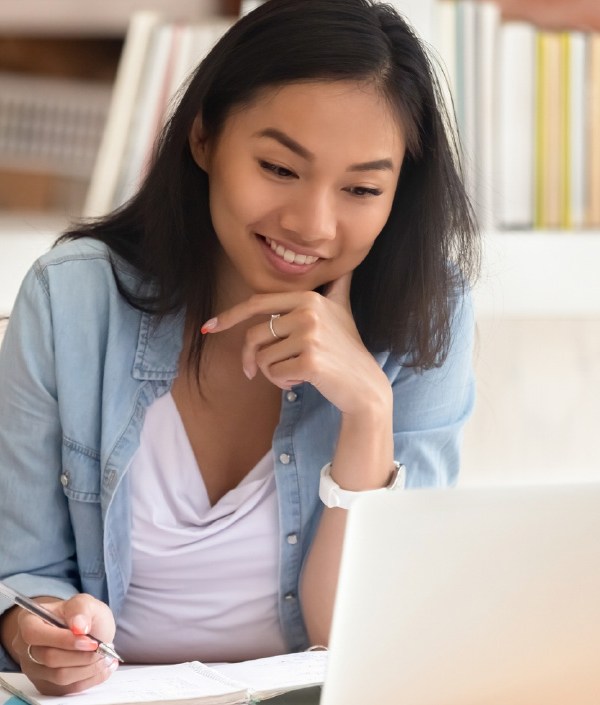 A woman sits in front of her laptop with a notebook open while taking notes, indicating she's learning how to write a first job resume.