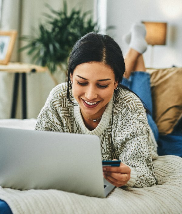 A woman lays on her bed while using her computer and holding her credit card, indicating she just learned about how credit cards work.