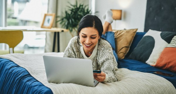 A woman lays on her bed while using her computer and holding her credit card, indicating she just learned about how credit cards work.