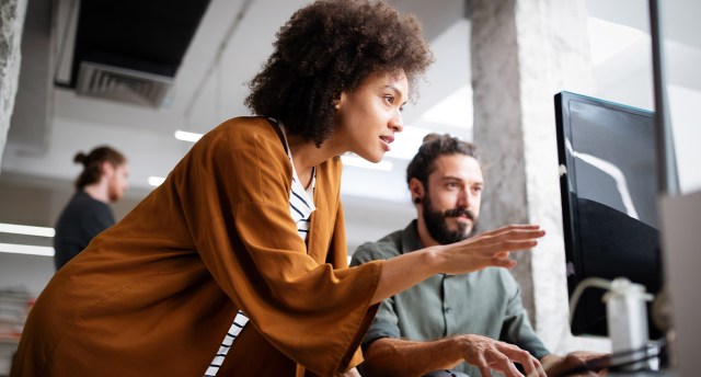 An IT manager, one of the most fulfilling jobs, points at the computer screen and guides her cowoker through a project.