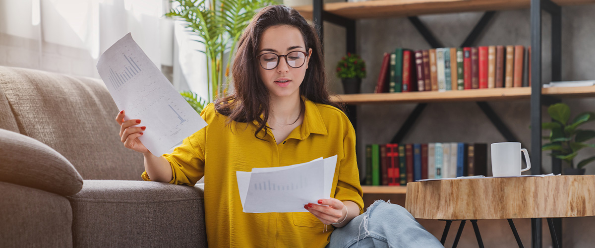 A woman in a yellow blouse sits on the floor of her living room reviewing documents, indicating she might be researching student loan forbearance.