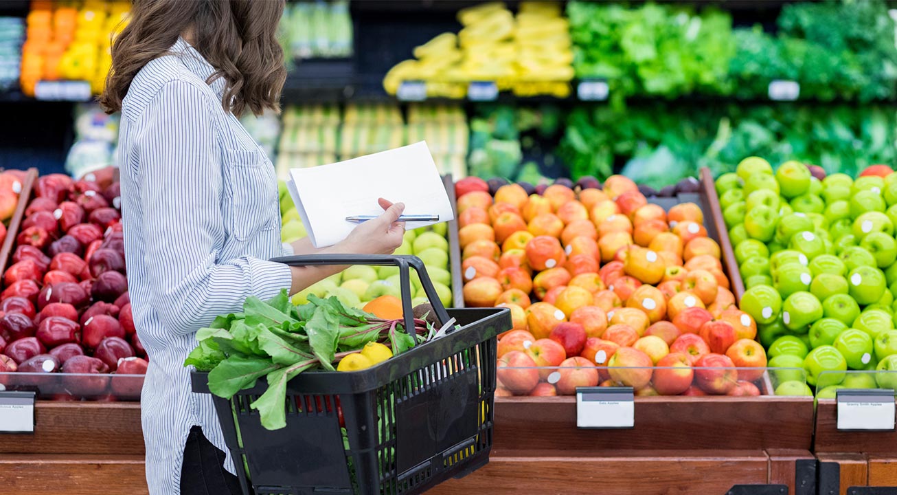 Person shopping in a grocery store with a basket of fruits and veggies looking at a row of assorted apples in open bins.